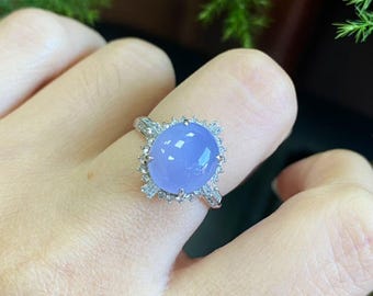 May include: A silver ring featuring a large, oval-shaped, light blue gemstone. The gemstone is surrounded by a halo of smaller, clear stones. The ring is on a finger.