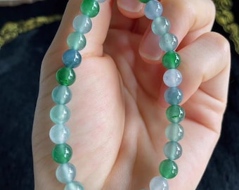 May include: A beaded bracelet featuring a variety of green and blue-grey gemstones. The bracelet is strung on a thin, clear cord and is held in a hand. The beads are round and vary in color, creating a gradient effect.
