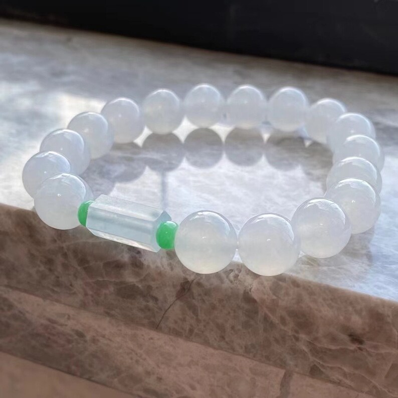 May include: A white jade bracelet with a green jade accent. The bracelet is made of round beads and has a rectangular green jade bead in the center.