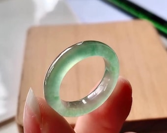 May include: A translucent jade ring with a smooth, polished surface. The ring is a light green color and appears to be made of a single piece of jade. The ring is held up by a person's fingers.
