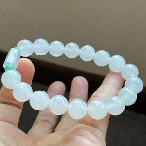 May include: A white jade bracelet with a green jade bead accent. The bracelet is made of round beads and is strung on a clear elastic cord.