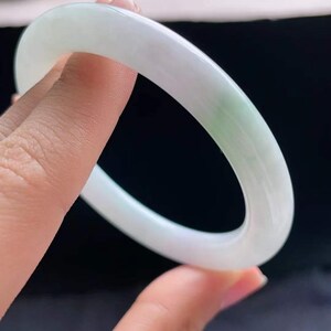 Op de afbeelding: Een doorschijnende jade armband. De armband is voornamelijk wit met subtiele groene adering. Het gladde, gepolijste oppervlak reflecteert licht en benadrukt de natuurlijke variaties in de steen. De armband wordt tegen een donkere achtergrond gehouden.