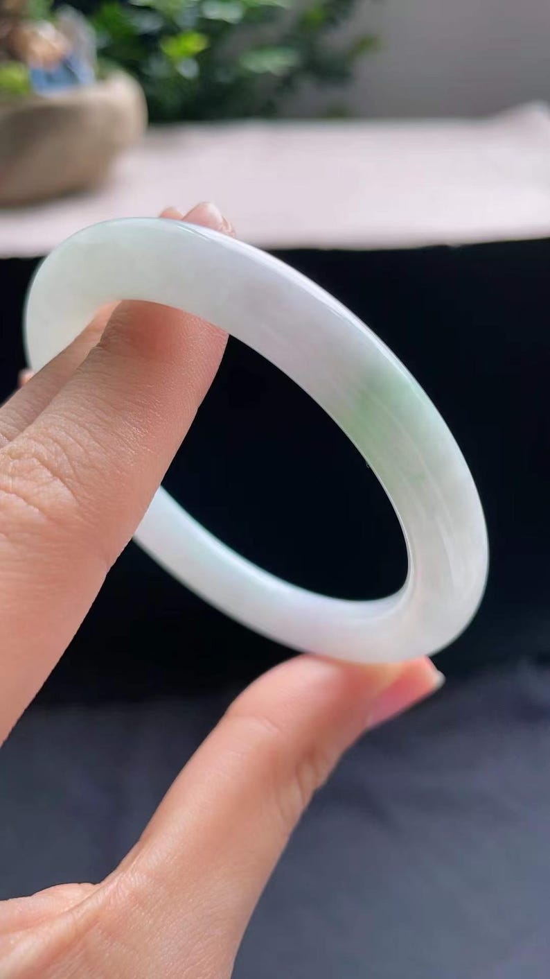 Op de afbeelding: Een doorschijnende jade armband. De armband is voornamelijk wit met subtiele groene adering. Het gladde, gepolijste oppervlak reflecteert licht en benadrukt de natuurlijke variaties in de steen. De armband wordt tegen een donkere achtergrond gehouden.