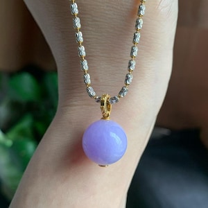 May include: A necklace with a spherical, lavender-coloured pendant. The pendant hangs from a gold-toned bail and a silver and gold-toned chain. The necklace is shown against a neutral background.