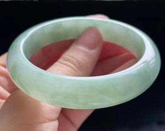 May include: A pale green jade bangle. The polished surface of the bracelet reflects light, highlighting the natural colour variations. The jade has a translucent quality. A classic piece of jewellery.