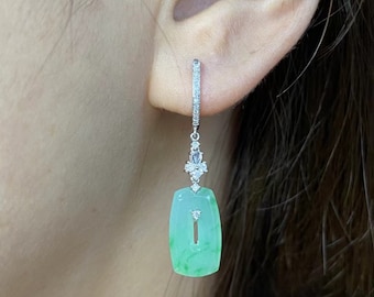 May include: A close-up of a single earring. The earring features a rectangular, light green jade pendant with a vertical slit. The pendant is suspended from a diamond-studded bar and a cluster of diamonds. The earring is worn on an ear.