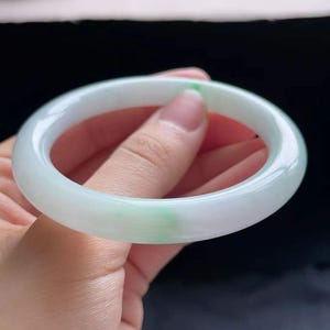 Op de afbeelding: Een jade armband met een glad, gepolijst oppervlak. De armband heeft een lichtgroene en witte kleur, met een doorschijnend uiterlijk. De jade is een natuursteen en de armband is een klassiek sieraad.