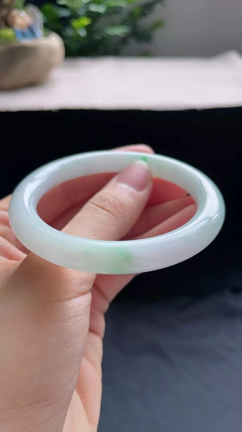 Op de afbeelding: Een jade armband met een glad, gepolijst oppervlak. De armband heeft een lichtgroene en witte kleur, met een doorschijnend uiterlijk. De jade is een natuursteen en de armband is een klassiek sieraad.