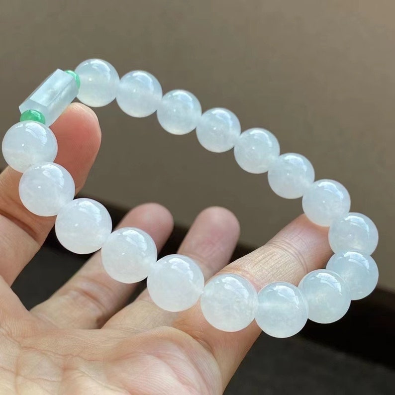 May include: A white jadeite bracelet with a green jadeite bead accent. The bracelet is made of round beads and is approximately 7 inches in length.