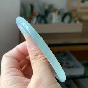 Op de afbeelding: Een lichtblauwe jade armband. Het gladde, gepolijste oppervlak reflecteert licht en toont de doorschijnende kwaliteit van de steen. De armband is cirkelvormig, ontworpen om om de pols te dragen. De kleur is een zacht, lichtblauwgroen.