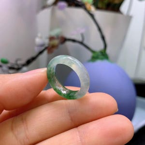 May include: A jade ring with a translucent appearance, showcasing a blend of green and white hues. The ring is held in a hand, with a blurred background featuring a purple vase and greenery. The ring's smooth surface reflects light.