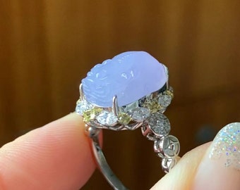 May include: A lavender jade ring with a carved design and a halo of diamonds and yellow gemstones. The ring is set in silver.
