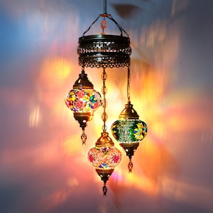 May include: A hanging chandelier with three colorful mosaic glass lamps. The lamps are in shades of red, green, and yellow, with intricate patterns. The metal frame has a bronze finish, and the fixture hangs from a chain.