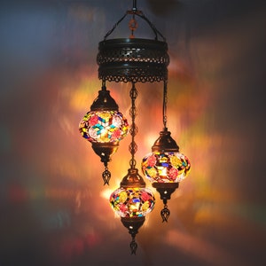 Puede incluir: Una lámpara de araña de tres luces con pantallas de vidrio de mosaico de colores. Las pantallas son redondas y presentan una variedad de colores, incluyendo rojo, azul, verde y amarillo. La lámpara de araña está hecha de metal y tiene un acabado marrón oscuro.