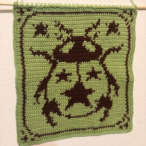 May include: A square, handmade crochet wall hanging in shades of green and brown. The design features a central star shape surrounded by other star shapes and decorative elements. The piece is hung from a wooden dowel.