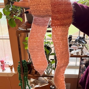 Op de afbeelding: Een handgemaakte gehaakte sjaal in tinten perzik, roze en lichtpaars. De sjaal heeft een getextureerd, pluizig uiterlijk met een uniek ontwerp. De sjaal wordt voor een raam gehouden.