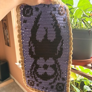 May include: Handmade crochet wall hanging featuring a purple and black design with a brown border. The design appears to be a stylized face or creature. The piece is held up in front of a window with plants.