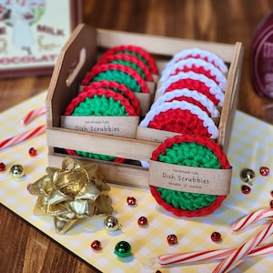 May include: A wooden crate filled with handmade tulle dish scrubbies. The scrubbies are red, green, and white, with a circular design. The text "Dish Scrubbies" is printed on a brown label. A gold bow and Christmas decorations are in the foreground.