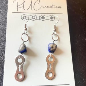 Peut inclure: Boucles d'oreilles faites à la main avec des maillons de chaîne de vélo argentés et de petites pierres bleu-gris. Les boucles d'oreilles sont suspendues à des crochets argentés. La carte de support affiche le texte "RUC creations" et le compte Instagram "@r.u.c.creations".