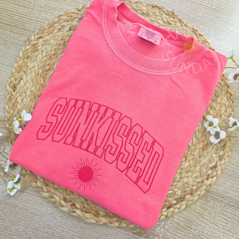 May include: A coral-colored t-shirt with the word "SUNKISSED" in a bold, arched font. Below the text is a sun graphic. The shirt is folded on a woven mat, with small white flowers and greenery in the background.