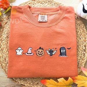 Könnte beinhalten: Ein orangefarbenes T-Shirt mit Rundhalsausschnitt und gestickten Halloween-Motiven, darunter ein weißer Geist, ein rosa Hexenhut, ein orangefarbener Kürbis, eine graue Fledermaus und ein schwarzer Grabstein mit der Aufschrift RIP. Comfort Colors Etikett.