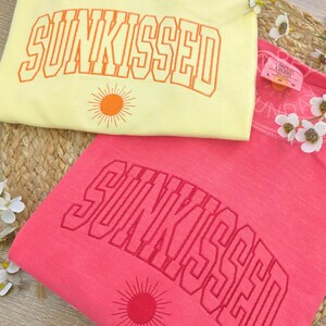 May include: Two sweatshirts, one yellow and one pink, with the word "SUNKISSED" embroidered across the front in a matching color. Each shirt has a sun graphic below the text. The yellow shirt has orange embroidery, and the pink shirt has red embroidery.