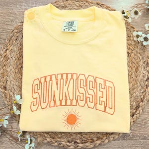 May include: A light yellow sweatshirt with the word "SUNKISSED" embroidered in orange block letters. A small orange sun graphic is below the text. The sweatshirt is folded on a woven mat.