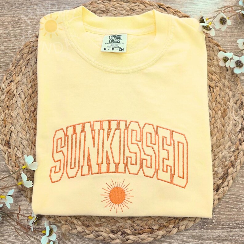 May include: A light yellow sweatshirt with the word "SUNKISSED" embroidered in orange block letters. A small orange sun graphic is below the text. The sweatshirt is folded on a woven mat.