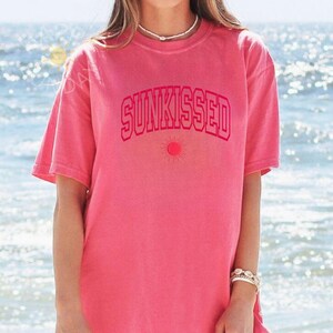 May include: A pink t-shirt with the word "SUNKISSED" in a matching pink, arched font. Below the text is a small sun graphic. The shirt has short sleeves and a crew neck. The background is a blurred view of the ocean.