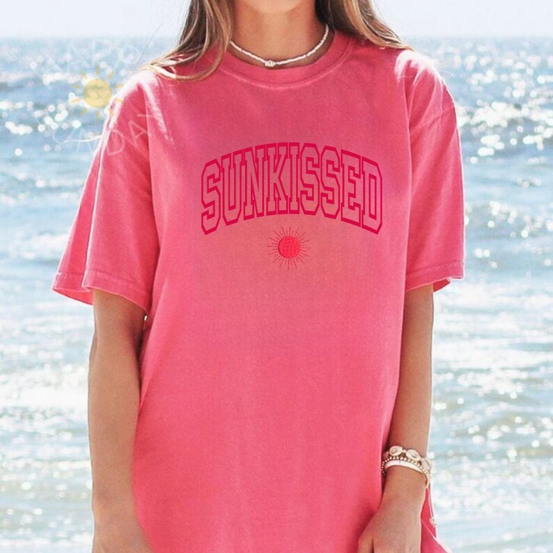 May include: A pink t-shirt with the word "SUNKISSED" in a matching pink, arched font. Below the text is a small sun graphic. The shirt has short sleeves and a crew neck. The background is a blurred view of the ocean.