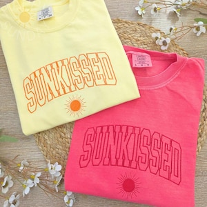 May include: Two t-shirts, one yellow and one pink, with the word "SUNKISSED" in orange lettering. Each shirt has a sun graphic below the text. The shirts are folded on a woven mat.