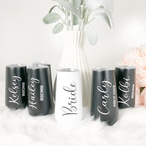 May include: Six personalized stainless steel tumblers with black and white lettering. The tumblers are arranged in a row on a white fluffy surface. The tumblers read "Bride", "Maid of Honor", "Carly", "Kolbi", "Hailey", and "Kelsey".