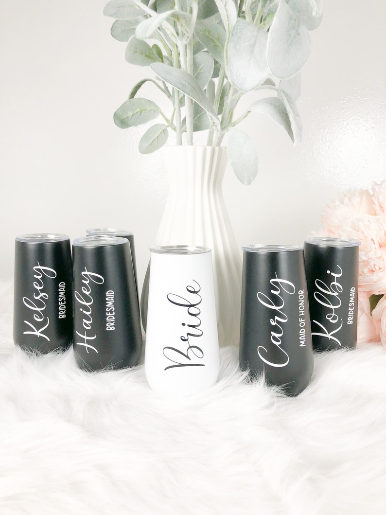 May include: Six personalized stainless steel tumblers with black and white lettering. The tumblers are arranged in a row on a white fluffy surface. The tumblers read "Bride", "Maid of Honor", "Carly", "Kolbi", "Hailey", and "Kelsey".