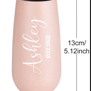 May include: A pink stainless steel champagne flute with the name "Ashley" and the word "Bridesmaid" in white lettering. The flute is 5.12 inches tall and 2.36 inches wide.