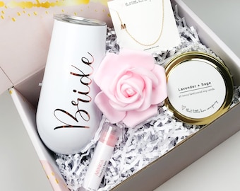 May include: A white gift box filled with a white tumbler with the word "Bride" in rose gold lettering, a pink rose, a small lip balm tube, a gold candle with the text "Lavender + Sage" and a gold necklace with a letter "M" charm.