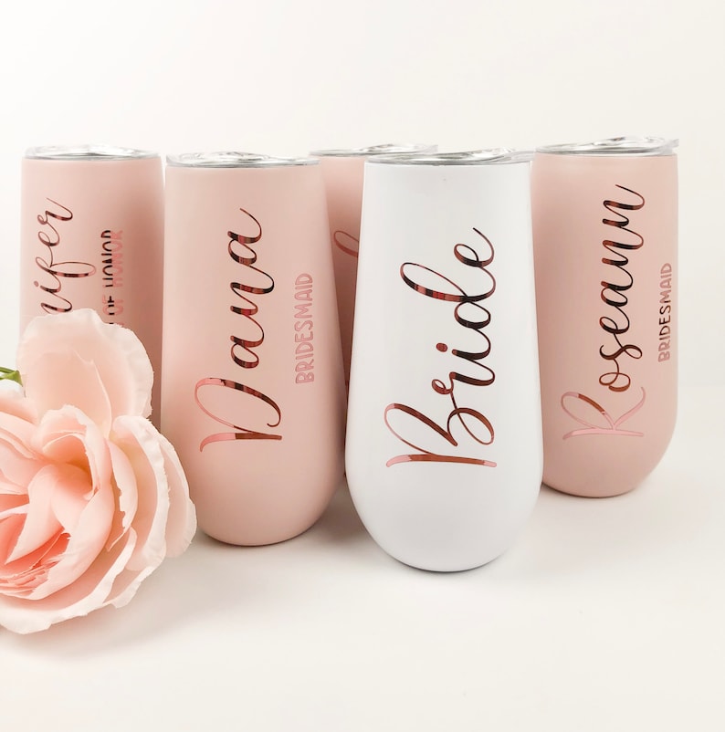May include: Set of five stainless steel champagne flutes with rose gold lettering. The flutes are in two colors: pink and white. The text on the flutes reads "Bride", "Wife", "Nana Bridesmaid", "Roseann Bridesmaid".