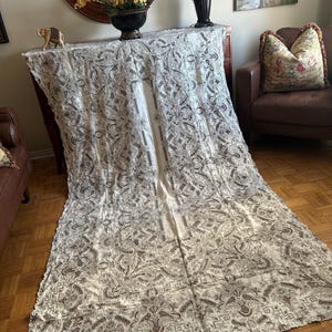 May include: A cream-coloured tablecloth with a paisley pattern and a scalloped edge. The intricate design features brown and grey floral and geometric elements. The tablecloth is draped over a dark wooden table, creating a vintage aesthetic.