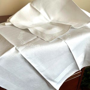Puede incluir: Mantel de lino blanco y servilleta plegada a juego. El mantel presenta un sutil estampado floral y un monograma en la esquina. La servilleta está doblada sobre el mantel. Los artículos están sobre una superficie de madera oscura.