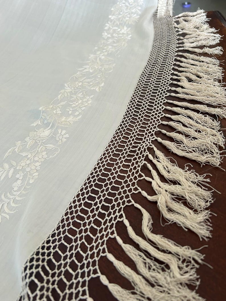K&ouml;nnte beinhalten: Nahaufnahme eines wei&szlig;en Textils mit floraler Stickerei und geh&auml;keltem Rand. Der Rand weist ein geometrisches Muster und lange Fransen auf. Der Stoff scheint leicht und halbtransparent zu sein, mit einer zarten Textur.