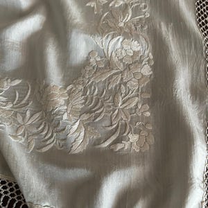 K&ouml;nnte beinhalten: Ein hellblaues Textil, m&ouml;glicherweise Seide, mit detaillierter floraler Stickerei in einem helleren Farbton. Der Rand hat eine zarte, offene H&auml;kelborte. Die Stickerei konzentriert sich in der Ecke und zeigt detaillierte Blumen- und Blattmuster.