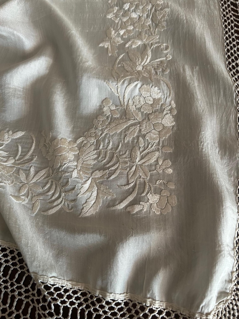 K&ouml;nnte beinhalten: Ein hellblaues Textil, m&ouml;glicherweise Seide, mit detaillierter floraler Stickerei in einem helleren Farbton. Der Rand hat eine zarte, offene H&auml;kelborte. Die Stickerei konzentriert sich in der Ecke und zeigt detaillierte Blumen- und Blattmuster.