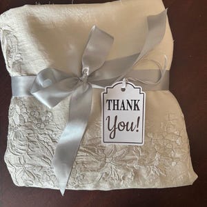 K&ouml;nnte beinhalten: Ein gefaltetes, cremefarbenes Stoffst&uuml;ck mit floraler Stickerei ist mit einem silbernen Band und einer Schleife gebunden. Ein Etikett mit den Worten "Thank You!" ist angebracht. Der Stoff scheint ein dekorativer Artikel zu sein.