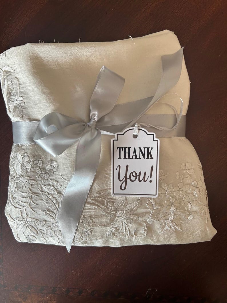 K&ouml;nnte beinhalten: Ein gefaltetes, cremefarbenes Stoffst&uuml;ck mit floraler Stickerei ist mit einem silbernen Band und einer Schleife gebunden. Ein Etikett mit den Worten "Thank You!" ist angebracht. Der Stoff scheint ein dekorativer Artikel zu sein.