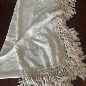 K&ouml;nnte beinhalten: Cremefarbenes rechteckiges Textil mit floraler Stickerei und Fransen. Das Textil weist detaillierte Blumenmuster auf, die entlang der Mitte und der Kanten gestickt sind. Die Kanten sind mit einer gewebten Bord&uuml;re und langen, flie&szlig;enden Fransen versehen.