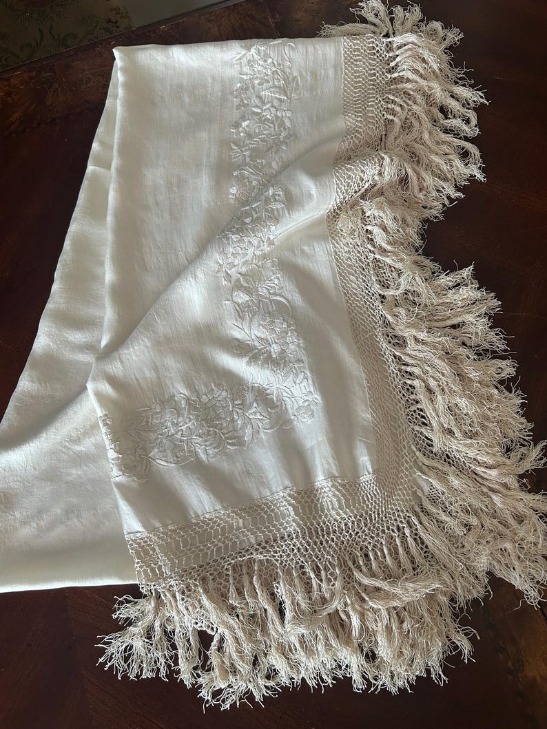 K&ouml;nnte beinhalten: Cremefarbenes rechteckiges Textil mit floraler Stickerei und Fransen. Das Textil weist detaillierte Blumenmuster auf, die entlang der Mitte und der Kanten gestickt sind. Die Kanten sind mit einer gewebten Bord&uuml;re und langen, flie&szlig;enden Fransen versehen.