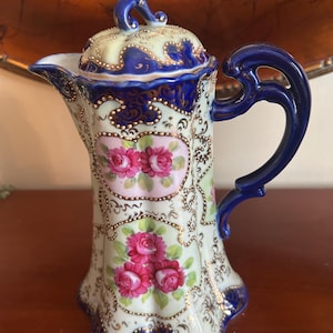 Puede incluir: Una cafetera de porcelana vintage con tapa, con una base color crema con detalles azules y dorados. La cafetera está adornada con diseños florales de rosas rosas y un asa azul.