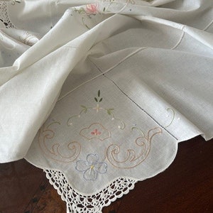 Może przedstawiać: Biały obrus z delikatnym haftem kwiatowym i koronkowym brzegiem. Obrus jest złożony na pół, odsłaniając skomplikowane detale haftu.