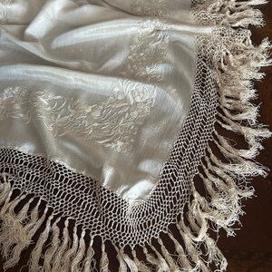 K&ouml;nnte beinhalten: Cremefarbener Stoff mit floraler Stickerei und geh&auml;keltem Fransenrand. Der Stoff ist drapiert und zeigt die Textur und das Design. Die Fransen sind lang und verleihen dem Textil ein dekoratives Element.