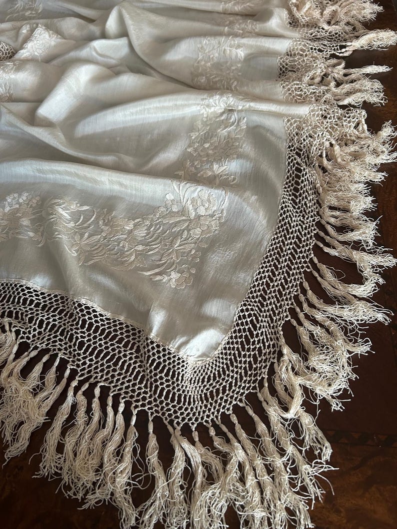 K&ouml;nnte beinhalten: Cremefarbener Stoff mit floraler Stickerei und geh&auml;keltem Fransenrand. Der Stoff ist drapiert und zeigt die Textur und das Design. Die Fransen sind lang und verleihen dem Textil ein dekoratives Element.