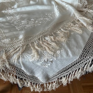 K&ouml;nnte beinhalten: Eine cremefarbene, runde Tischdecke mit aufwendiger Blumenstickerei und einem geh&auml;kelten Rand. Der Rand ist mit langen, zarten Fransen versehen. Der Stoff scheint ein weiches, flie&szlig;endes Material zu sein.